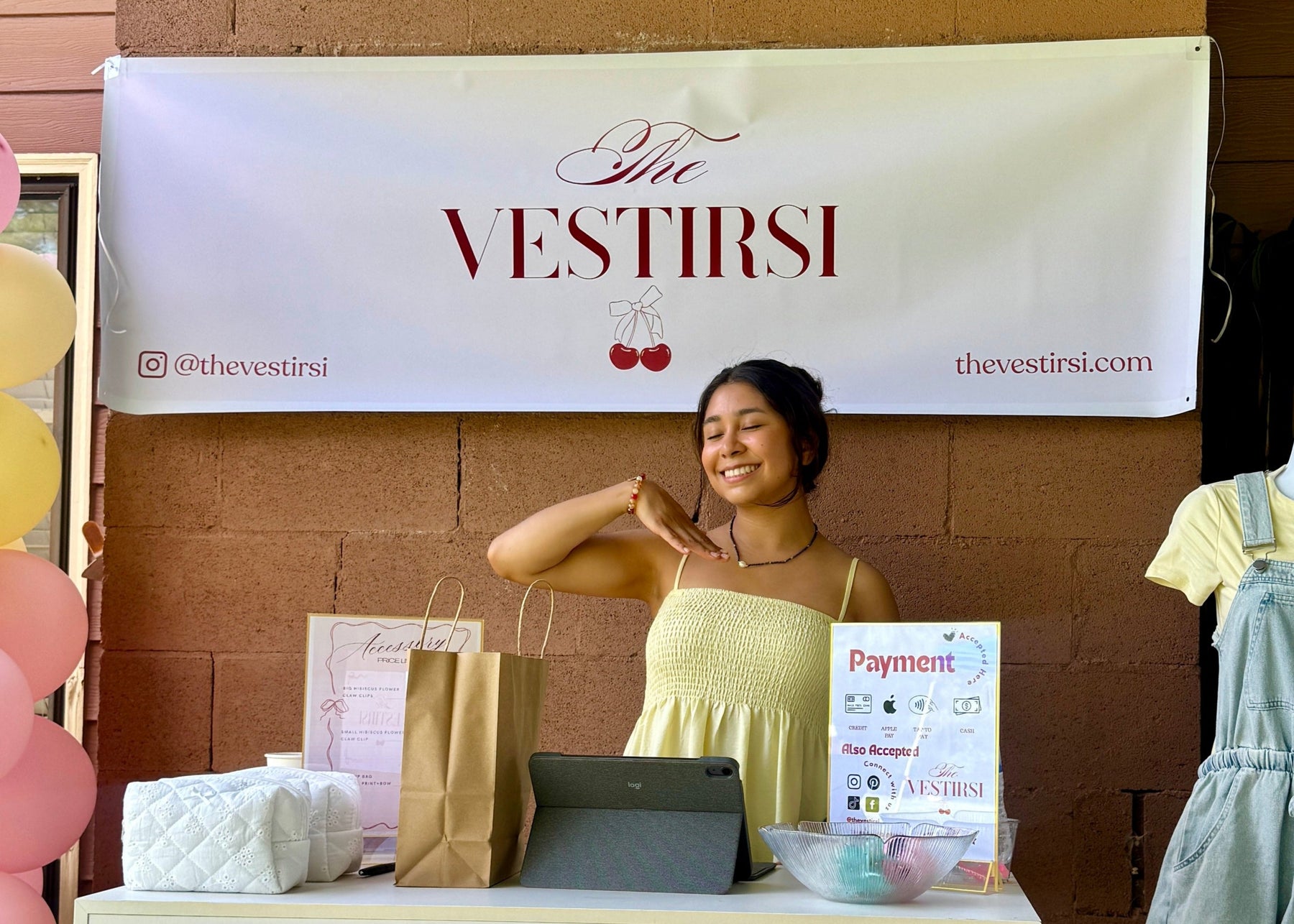 Woman standing behind a table with promotional materials and dresses, with 'The Vestirsi' brand banner in the background.